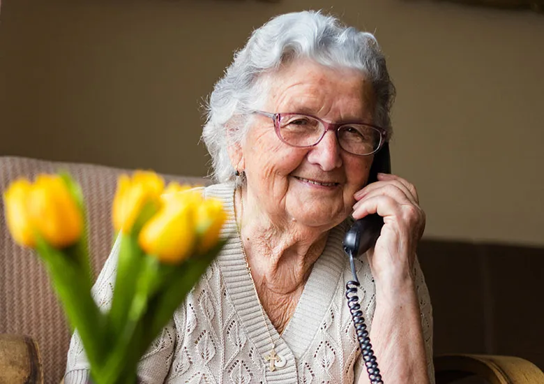 An elderly woman with glasses smiles while talking on a corded phone, with yellow tulips blurred in the foreground.