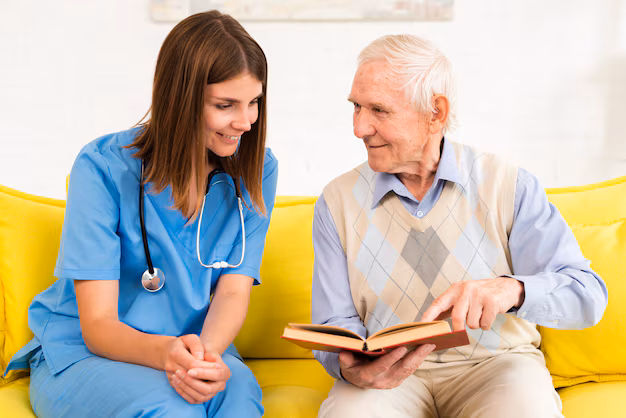 A healthcare professional in blue scrubs sits with an older man on a couch as he shows her an open book.